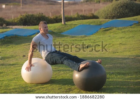 Fitness coach balancing himself on top of 2 fit-balls as part of an exercise. Skating pump track in the background.