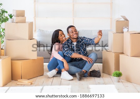 Full length portrait of beautiful black couple taking selfie on floor on moving day, surrounded by cardboard boxes. Happy young guy and his pretty wife making photo of themselves while relocating