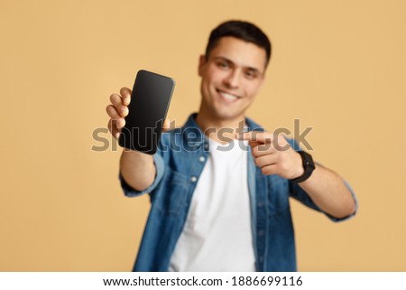 It is cool, best mobile app and digital ad. Happy young man in casual shows finger at smartphone with blank screen, selective focus on device, isolated on beige background, studio shot, free space