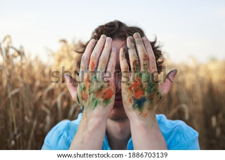 Close-up picture of male artist covering his face with stained with paint hands on wheat field in summer.Painting workshop in rural countryside.Artistic education concept. Outdoors leisure activities.