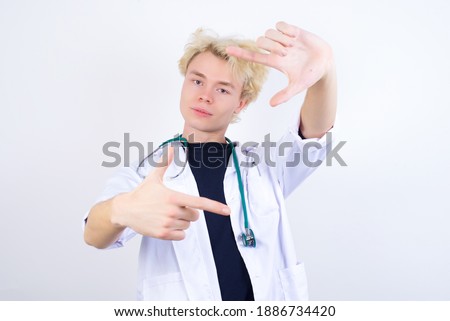 Positive young handsome Caucasian doctor man standing against white background with cheerful expression, has good mood, gestures finger frame actively at camera.