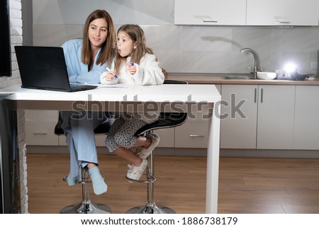 Cheerful smiling happy mom and her nosy little daughter sit at the table in the kitchen at home and use laptop to watch cartoons, on weekends