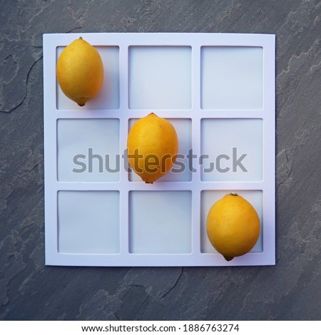Four real yellow lemons in a diagonal row on a white geometric square grid. Noughts and crosses. Grey slate background. Simple elegant minimal flat lay from above.