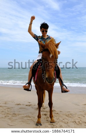 Asian tourist raise arm, horseback riding brown horse at Hua Hin beach, Prachuap Khiri Khan, Thailand