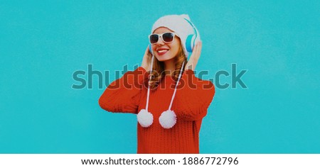 Winter portrait of happy smiling woman in wireless headphones listening to music wearing a red knitted sweater and white hat with pom pom on a blue background