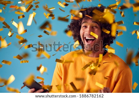 Celebrating Young Man With Mobile Phone Winning Prize And Showered With Gold Confetti In Studio Royalty-Free Stock Photo #1886782162
