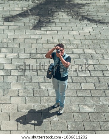 Curly hair boy dressed in denim, is taking pictures on a summer day, in the background a concrete floor and the shadow of a palm tree.