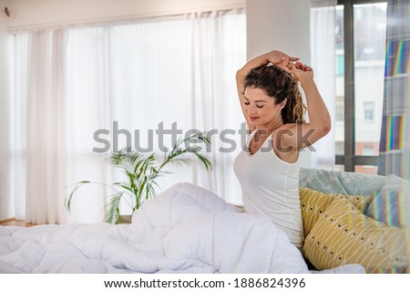 What a wonderful morning. Attractive young woman is stretching arms after sleeping. She is sitting on bed and smiling. Woman stretching in bed after waking up. It's a good morning to me