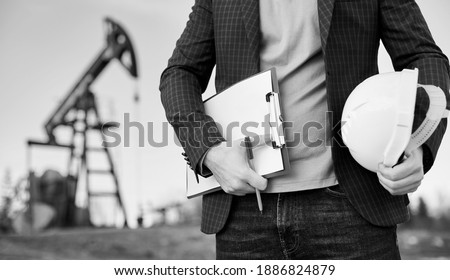 Cropped close-up snapshot of a man engineer holding his note and a pen in one hand and a white helmet in the other, standing in oil field, oil pump jack is on background. Black and white image