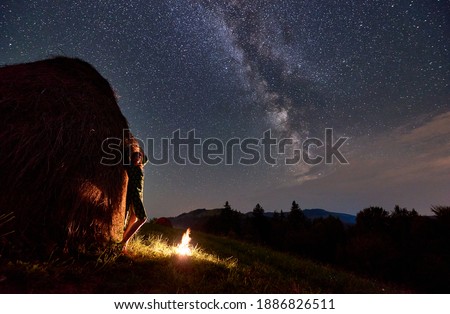Silhouettes of girls near a haystack and a bonfire against the backdrop of a valley of hills, under incredibly beautiful starry sky on which the Milky Way is visible at night