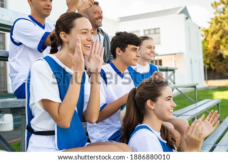 Players cheering for school football team sitting on stands. High school guys and girls clapping hands while watching sport match. Group of fans watching a sports event at stadium while encouraging. Royalty-Free Stock Photo #1886880010