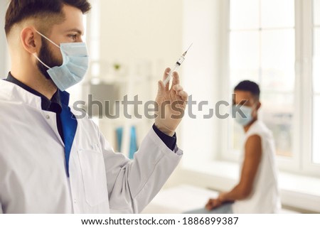 Closeup side view young male doctor in medical face mask preparing syringe for administering flu, AIDS or COVID-19 vaccine to patient against blurred hospital background with man waiting for injection