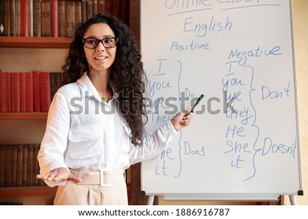 Foreign Language. Portrait of confident smiling young female lecturer wearing glasses teaching English pointing at grammar rules on board with marker, looking back at students at classroom Royalty-Free Stock Photo #1886916787
