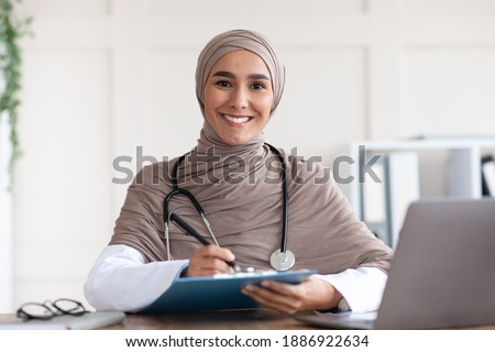 Beautiful muslim woman doctor writing in medical chart and smiling at camera, wearing hijab, sitting at workdesk with modern laptop in her cabinet in clinic or hospital, taking anamnesis, copy space