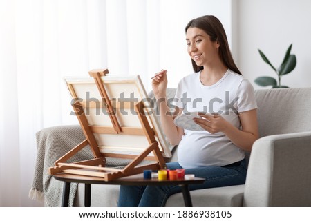 Painting While Pregnant. Happy Young Expecting Woman Drawing Picture On Easel At Home, Enjoying Art Therapy And Creative Leisure During Pregnancy, Young Awaiting Lady Sitting On Couch In Living Room
