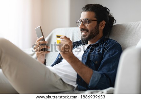 Cheerful Handsome Arab Guy Shopping Online With Smartphone And Credit Card At Home, Millennial Man With Glasses And Dental Braces Enjoying Rurchasing In Internet, Sitting On Couch In Living Room