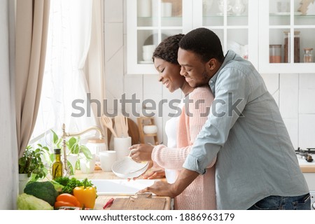 Romantic Black Couple Cleaning Dishes And Bonding In Kitchen, Making Domestic Chores Together. Loving African American Man Cuddling Wife From The Back, Enjoying Spending Time At Home, Side View Royalty-Free Stock Photo #1886963179