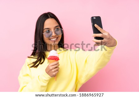 Young brunette girl holding a cornet ice cream over isolated pink background making a selfie
