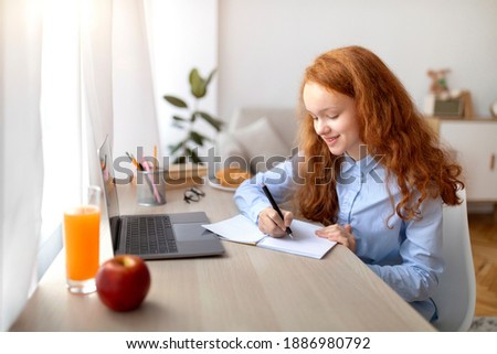 Remote Education Concept. Smiling young teenage girl sitting at table, using laptop and writing in her notebook or diary. Schoolgirl watching online course, taking notes, doing homework Royalty-Free Stock Photo #1886980792