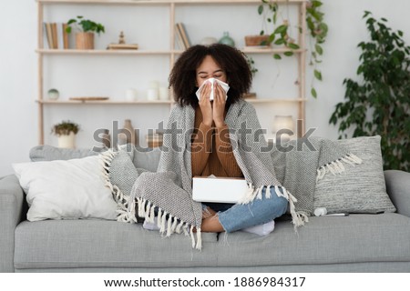 Sick day at home, treatment, unhealthy african american millennial woman has runny nose, cough and cold. Portrait of beautiful lady caught flu Illness covered with blanket on couch with napkins Royalty-Free Stock Photo #1886984317