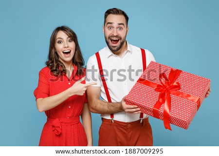 Excited young couple friends man woman in white red clothes pointing index finger on present box with gift ribbon bow isolated on blue background. Valentine's Day Women's Day birthday holiday concept