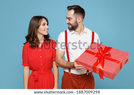 Smiling young couple friends man woman in white red clothes hold present box with gift ribbon bow look at each other isolated on blue background. Valentine's Day Women's Day birthday holiday concept