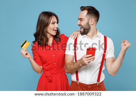 Happy young couple two friends man woman wearing white red clothes using mobile cell phone hold credit bank card doing winner gesture isolated on blue background. St. Valentine's Day holiday concept