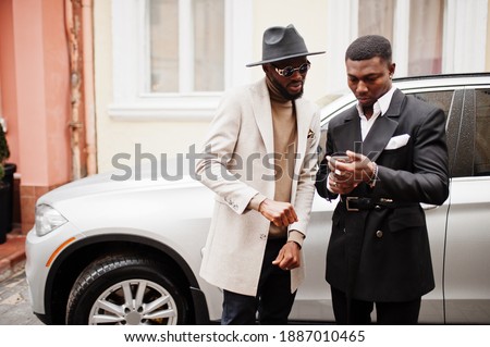 Two fashion black men stand near business car and look at cell phone. Fashionable portrait of african american male models. Wear suit, coat and hat. Royalty-Free Stock Photo #1887010465