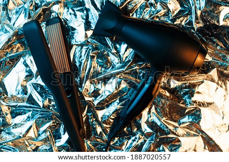 Hair dryer and hair straightener in neon lights on the foil with shiny crumpled surface background. 80s and 90s style.