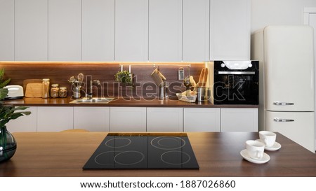 Panoramic view of contemporary interior with empty ceramic glass stove on wooden countertop against white kitchen cupboards, sink, electric oven and large refrigerator Royalty-Free Stock Photo #1887026860