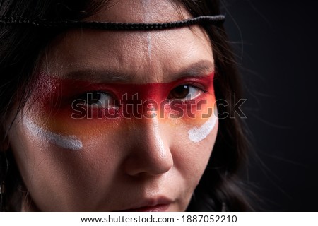 shamanic woman with warrior shaman make up isolated in studio, colourful paintings on her face and body, she is looking at camera, close-up eyes