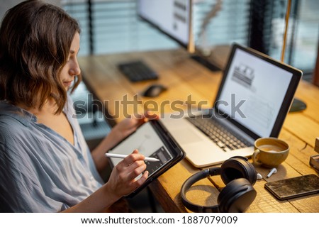 Young woman dressed casually having some creative work, drawing on a digital tablet, sitting at the cozy and stylish home office Royalty-Free Stock Photo #1887079009