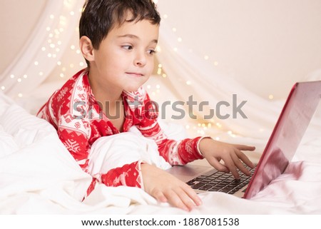 Happy child in red Christmas pajamas, playing on the Internet, watching cartoons, using a laptop, lies on a bed under a white canopy with lights in the bedroom. Winter vacation home concept.