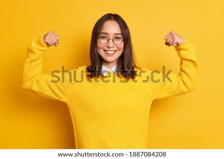 Studio shot of positive young Asian woman raises arms shows muscles pretends to be very strong and powerful smiles gently wears casual sweater isolated over yellow background. Look at my biceps Royalty-Free Stock Photo #1887084208