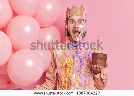 Excited young man smeared with cream holds chocolate cake wears paper crown on head carries inflated balloons enjoys celebration party event isolated over pink background. Happy birthday concept