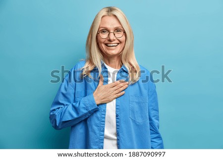 Sincere middle aged woman keeps hand on heart expresses gratitude smiles broadly being touched by heartleft words expresses truthy feelings dressed in fashionable clothing isolated over blue wall Royalty-Free Stock Photo #1887090997