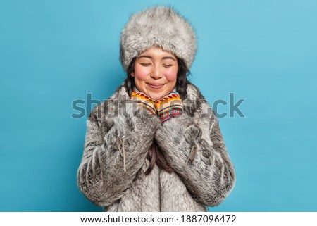 Dreamy satisfied young Asian woman closes eyes and smiles gently wears grey fur hat and coat has romantic mood poses against blue background. Eskimo female from north. Arctic aboriginal indoor