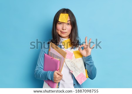 Everything is under control. Serious self confident young Asian woman makes okay gesture agrees to collaborate with colleague for preparing research work surrounded with papers and stickers.