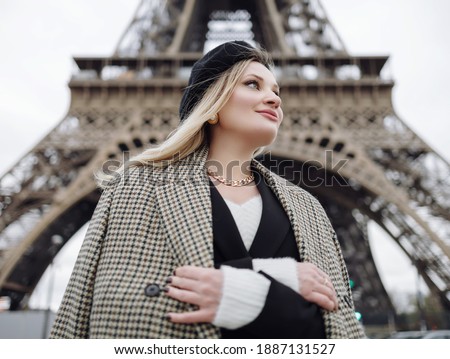 A stylishly dressed blonde stands at the foot of the Eiffel Tower in Paris. A girl wearing a jacket, black beret, vintage jewelry and a black bag. Royalty-Free Stock Photo #1887131527
