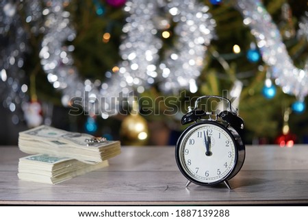 A small black alarm clock shows 12 o'clock, stands on a light table with a blurred background and bokeh lights. Christmas concept. Minutes about New year.  Midnight, midday.                        