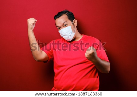 Young asian man wearing a medical face mask while raise his hand up to encourage and protects against the spread of corona virus disease. red background Royalty-Free Stock Photo #1887163360