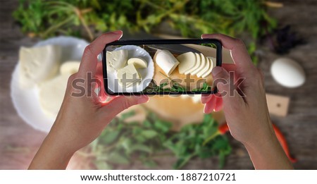Hands holding smartphone and taking photo of sliced cheese on a wooden table with green herbs