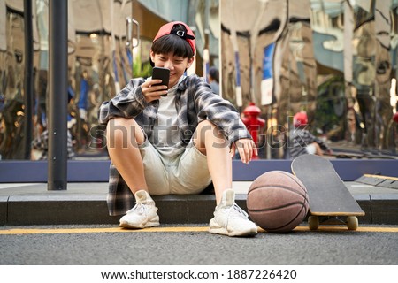 teenage asian kid with basketball and skateboard sitting on curb of street looking at cellphone