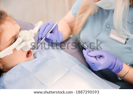 Female dentist checking child teeth with dental explorer and mirror while girl lying in dental chair with inhalation sedation. Concept of pediatric, sedation dentistry and dental care. Royalty-Free Stock Photo #1887290593