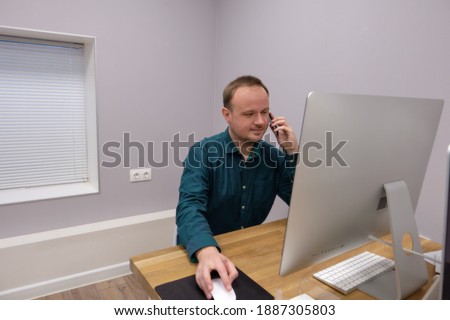A man in a dark green shirt sits in an office in front of a computer screen, looking into it. He holds the mouse with one hand, and the other phone near the ear.