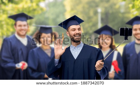 education, graduation and people concept - male graduate student in mortar board and bachelor gown taking picture with smartphone on selfie stick over international group at park on background