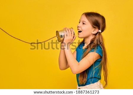 Positive teenage girl says in a tin can telephone, playing in retro connected chat, on yellow background. Crossed wires concept and telephone conversation Royalty-Free Stock Photo #1887361273