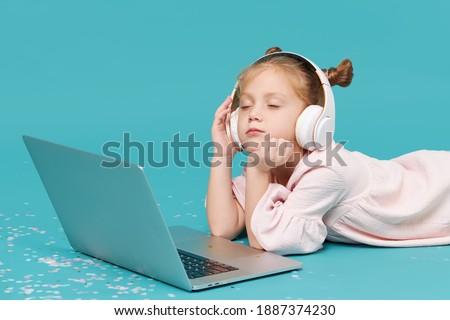 Smart child girl in wireless headphones using laptop on cyan background. Education, online study, tv, distance learning. Primary school student during quarantine. Educational games for children 