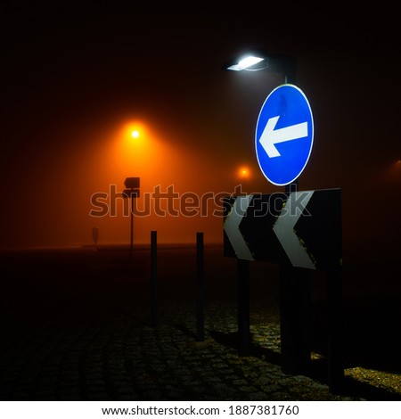 A blue road sign at night pointing to the left. There is also a sign post being illuminated by an orange street lamp which is obscured by fog, giving an atmospheric feeling to the picture.