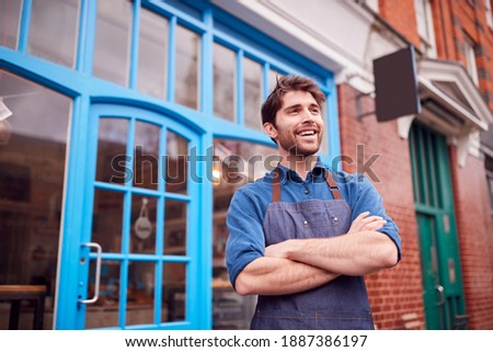 Smiling Male Small Business Owner Wearing Apron Standing Outside Shop On Local High Street Royalty-Free Stock Photo #1887386197